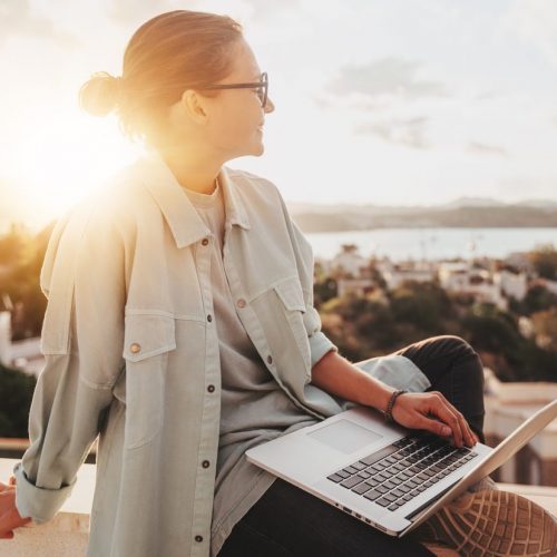 Beautiful curly young girl with glasses working on a laptop with a city view at sunset. Modern technologies and urban lifestyle. Freelance and online education